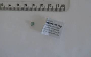 Fragment tabletki oksykodonu obok etykiety opakowania Oxypro 80 mg o przedłużonym uwalnianiu